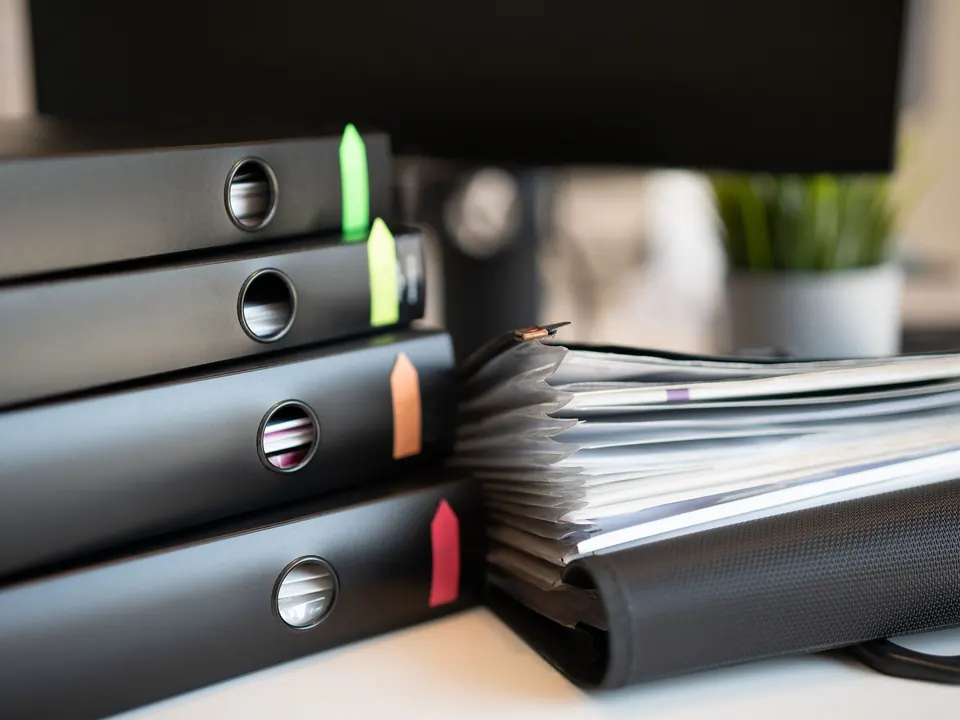 Stapel zwarte ordners met gekleurde tabs en een dossiermap op een bureau